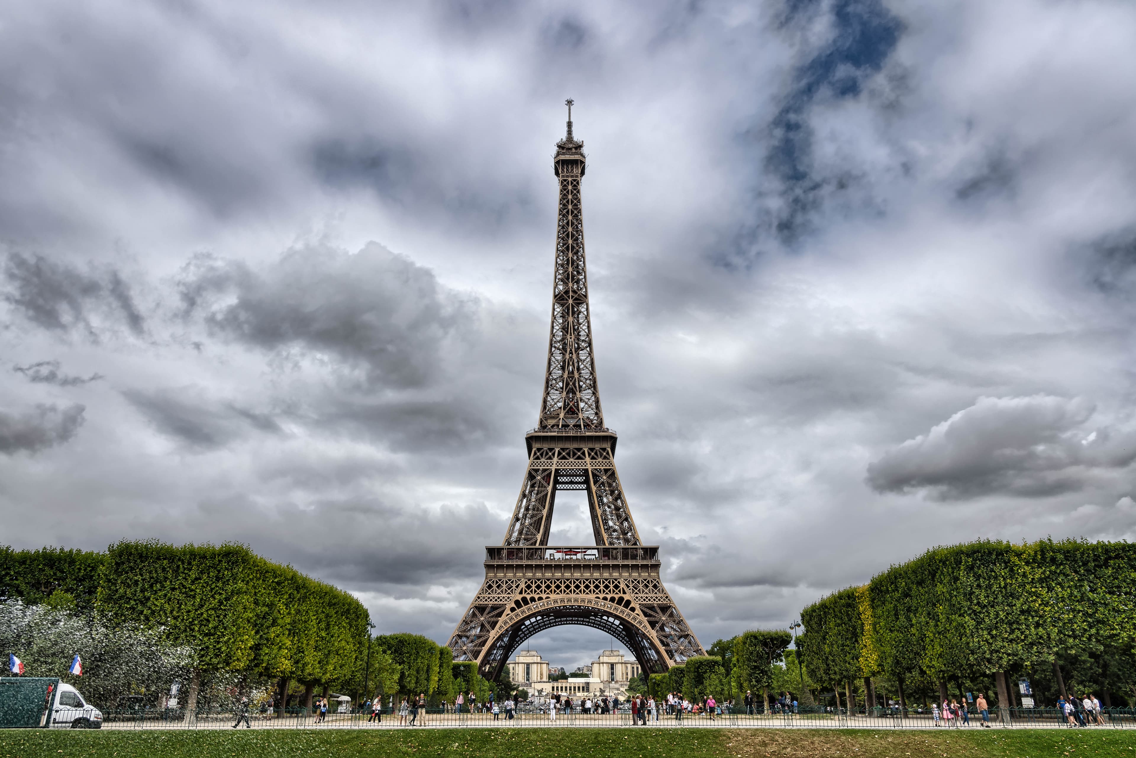 The Eiffel Tower centered between tree-lined paths of the Champ de Mars under dramatic clouds.